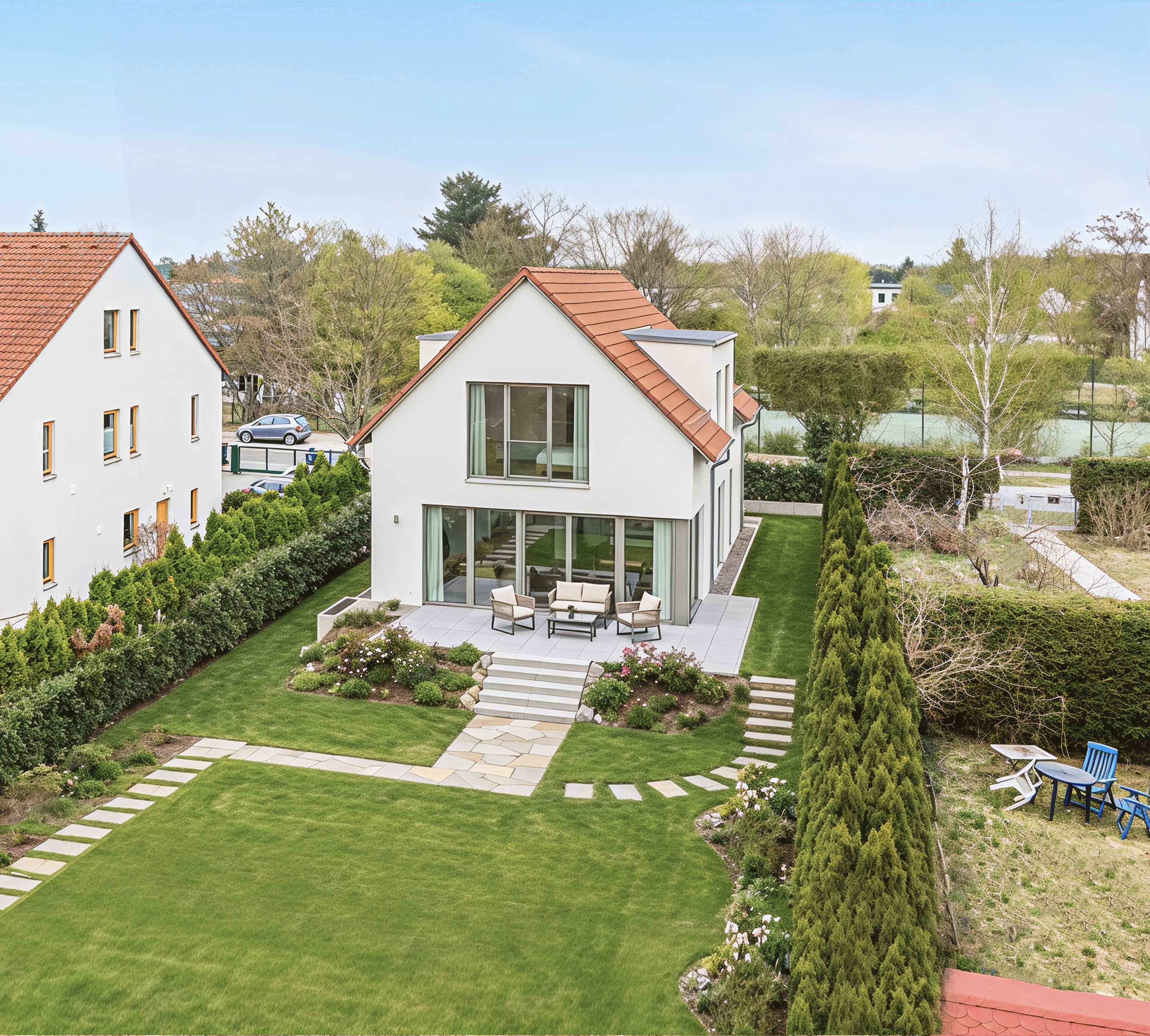 Nachher-Bild des virtuellen Außen-Stagings und professioneller Gartengestaltung eines modernen Einfamilienhauses. Der weitläufige Garten ist nun ein gepflegtes, gestagtes Paradies mit einem makellosen Rasen, sorgfältig angelegten Steinpfaden und einer möblierten Terrasse mit moderner Außenlounge-Garnitur. Der transformierte Zustand zeigt ein modernes Haus mit klaren Linien, großzügigen Glasfronten und einem neuen Ziegeldach, was zu einer bemerkenswerten Immobilienaufwertung führt. Das angrenzende Mehrfamilienhaus dient als Referenz.